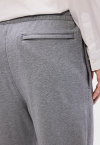 Pantalones de chándal grises con una textura suave, que cuentan con un solo bolsillo trasero y una cintura elástica. Se muestran de lado con una camiseta blanca.