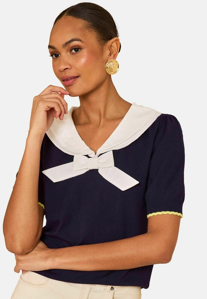 Hátrafésült hajú nő aranyérme-stílusú fülbevalóval, tengerészkék felsőben, fehér horgonyos gallérral és masnival, egyik kezét az arca közelében tartva pózol.