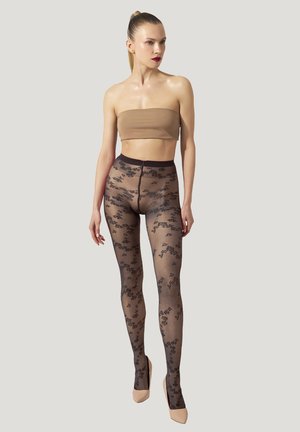 Donna con coda di cavallo bionda che indossa un top a fascia color nude, calze nere trasparenti con motivo floreale e tacchi alti beige, in piedi davanti a uno sfondo grigio chiaro.