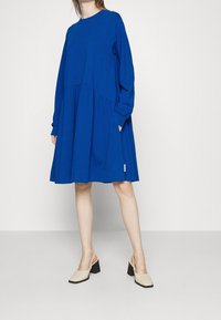 Sininen pitkähihaisen mekko, jossa on pyöreä pääntie, poimutettu vyötärö ja sivutaskut. Yhdistetty beigheihin lohkokorkoisiin slip-on kenkiin.