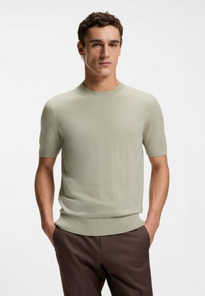 Uomo in piedi con le mani in tasca che indossa una maglia leggera beige a maniche corte e pantaloni marrone scuro su uno sfondo uniforme.