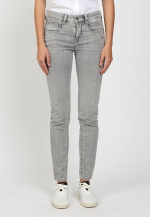 Slim-fit light gray jeans worn with white sneakers and a white tucked-in shirt, against a plain light background.