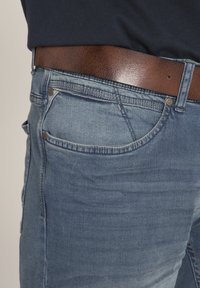Jeans in denim blu con una texture sbiadita, dotati di una cintura in pelle marrone. I jeans hanno tasche standard e dettagli con rivetti metallici.
