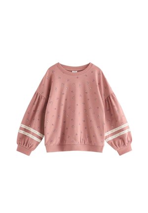 Smiling young girl with straight black hair wears a matching pink floral sweatshirt and pants with lace trim, standing against a plain background.