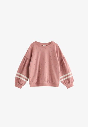 Jeune fille souriante aux cheveux noirs raides, portant un ensemble sweat-shirt et pantalon rose à fleurs avec bordure en dentelle, se tenant devant un fond uni.