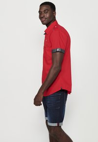 Camisa roja de manga corta con detalles de botones y acentos de denim en los puños enrollados, combinada con pantalones cortos de denim azul con dobladillos doblados.