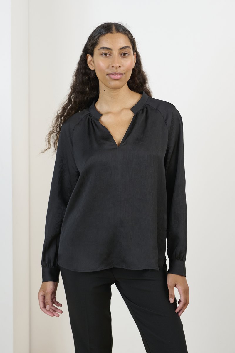 Blusa nera a maniche lunghe con scollatura a V, caratterizzata da una sottile texture a righe e dettagli arricciati sulle spalle. Abbinata a pantaloni neri.