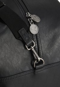 Nærbillede af sort lædertaske med lynlås, der har to runde Puma-logoer som trækstykker og en sort justerbar rem fastgjort med en metalclips.