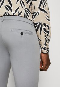 Sive krojene hlače z zadnjim žepom, ki ima črno gumb, v kombinaciji s patterned bluzo v bež, črni in beli listni zasnovi.