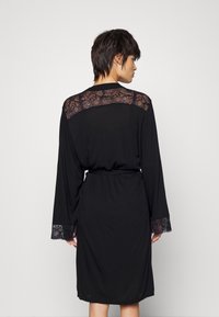 Robe noire à manches longues, accents en dentelle sur les épaules et les poignets, taille ceinturée, texture lisse et design à mi-genoux.