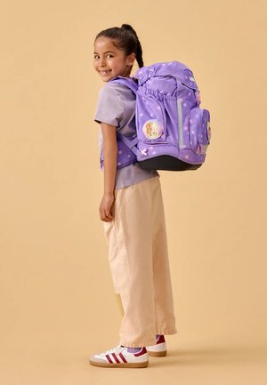 Girl smiling, standing sideways wearing a large purple polka-dot backpack, light purple shirt, beige pants, and white sneakers.