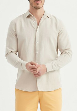 Homme portant une chemise beige à manches longues boutonnée et un pantalon jaune moutarde, debout les mains jointes à la taille devant un fond uni.