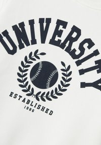 Baltā siltajā krekla ar melnu Druku. Teksts: "UNIVERSITĀTE" un "IZVEIDOTA 1986. GADĀ", ar beisbola grafiku un lauru lapu dizainu.