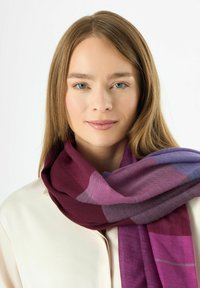 Écharpe violette et bordeaux avec une texture douce, présentant un motif géométrique et des bords à franges, portée lâchement autour du cou.