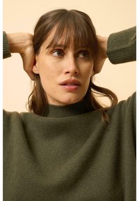 Pullover verde lavorato a maglia con colletto alto, morbida texture e dettagli a costine. Il modello ha i capelli di lunghezza media e uno sfondo neutro.