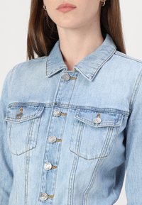 Svetlo modra denim jakna s ovratnikom, srebrnimi kovinskimi gumbi in dvema žepe na prsih z vidnimi šivi.