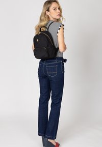 Svart ryggsäck med guldiga detaljer, dragkedjestängning och en logotyp. Modellen bär en randig topp och blå jeans, med röda plattformsskor.