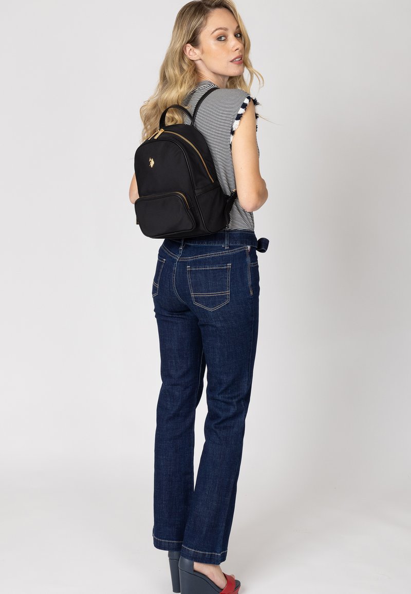 Svart ryggsäck med guldiga detaljer, dragkedjestängning och en logotyp. Modellen bär en randig topp och blå jeans, med röda plattformsskor.