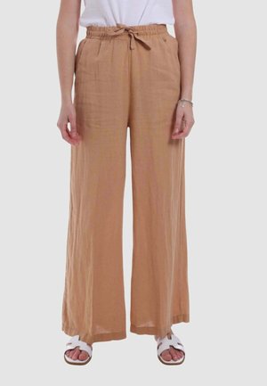 Persona che indossa pantaloni larghi marrone chiaro con coulisse in vita, sandali bianchi e top bianco, in piedi davanti a uno sfondo semplice.