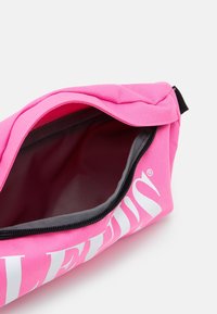 Różowa saszetka z zamkiem błyskawicznym o fakturowanej powierzchni, szarym wnętrzu i wyraźnym białym logo. Posiada górny zamek oraz małą pętlę do przyczepienia.