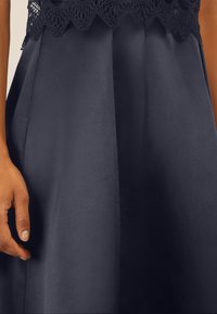 Abito blu navy con finitura satinata, caratterizzato da una sovrapposizione di pizzo sul corpetto e dettagli plissettati sulla gonna per texture e design.