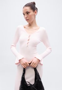 Ριμπ ύφασμα, μακρυμάνικη μπλούζα σε ροζ απόχρωση με V-λαιμόκοψη και διακοσμητικά κουμπιά. Το μοντέλο κρατά μια λαμπερή μαύρη τσάντα με καμπυλωτές λαβές.