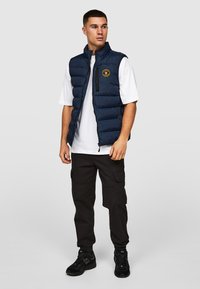 Marine blauwe puffer vest met horizontale stiksels, zij- en zakken met rits op de borst, en logo patch. Draag het over een wit T-shirt en combineer het met zwarte cargo broeken.