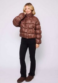 Brązowa kurtka puchowa z wysokim kołnierzem, błyszczącą teksturą i pikowanym wzorem. Połączona z czarnymi rozszerzanymi spodniami i brązowymi butami wsuwanymi.