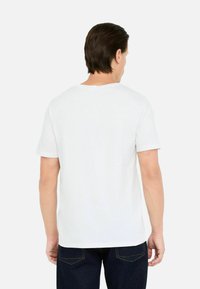 Mann mit kurzen dunklen Haaren, der ein schlichtes weißes T-Shirt mit kurzen Ärmeln und dunkelblaue Jeans trägt und mit dem Rücken zu einem hellen, einfarbigen Hintergrund steht.