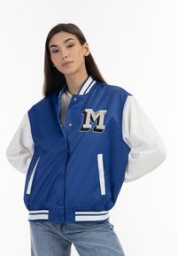 Giacca varsity in satin blu con maniche in pelle bianca, strisce bianche sui polsini e sull'orlo, con una patch "M" sul petto e chiusura con bottoni a pressione.