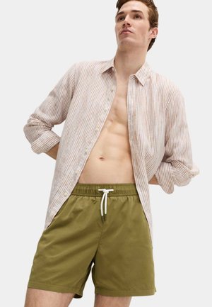 Ung mand iført en let, stribet skjorte åben over bare bryst og olivengrønne shorts med snøre, stående med hænderne bag ryggen.