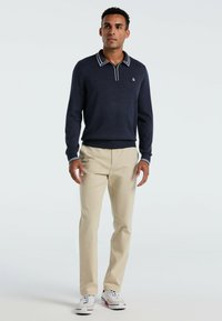 Marinefarbener Pullover mit weißem gestreiften Kragen, kombiniert mit beigen Hosen und weißen Sneakers mit roten Akzenten. Glatte Textur, lässige Passform.