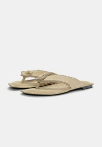 Beige platta sandaler med en tångrem och dekorativa metallnitar, tillverkade av mjukt mockamaterial, med en tunn sula.