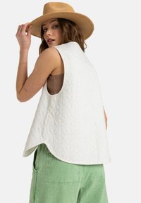 Chaleco blanco acolchado con un patrón floral, de corte holgado y dobladillo redondeado. Combinado con pantalones cortos de cargo verdes y un sombrero de paja.