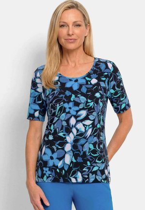Frau mit glatten blonden Haaren, die ein kurzärmeliges marineblaues Shirt mit blauen und weißen Blumenmustern und einfarbige blaue Hosen trägt.
