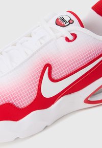Fehér és piros Nike Air Max sneaker piros kiegészítőkkel, fehér swoosh-al és textúrázott hálós felső résszel, közeli képen ábrázolva.