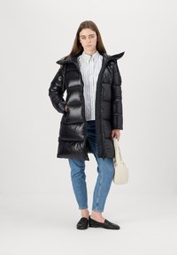 Zwarte pufferjas met een glanzende afwerking, voorzien van een ritssluiting aan de voorkant, hoge kraag en een oversized pasvorm. Gepaard met blauwe jeans en zwarte instappers.