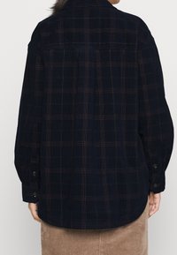 Camicia in flanella a quadri blu navy con maniche lunghe, polsini abbottonati e orlo arrotondato. Presenta un tessuto testurizzato e sottili accenti rossi nel motivo.
