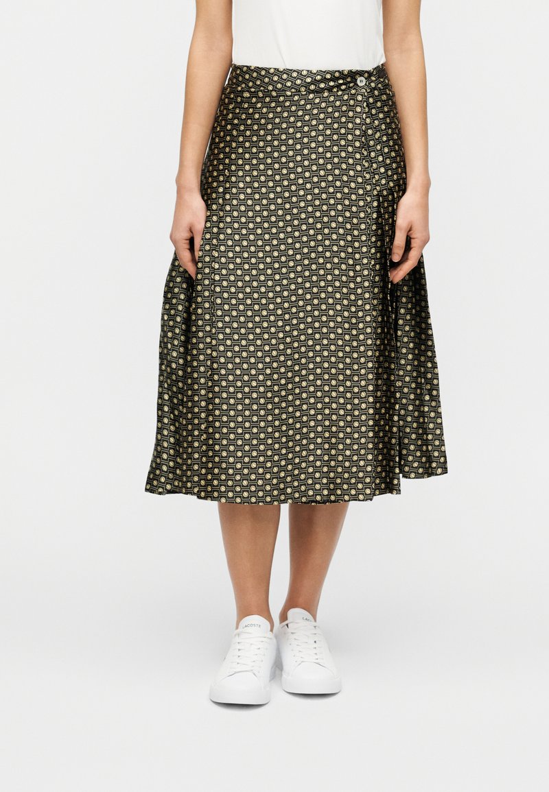 Μαύρη φούστα με σχέδιο γεωμετρικών μοτίβων, με υψηλή μέση και αέρινη σιλουέτα, συνδυασμένη με λευκά αθλητικά παπούτσια.