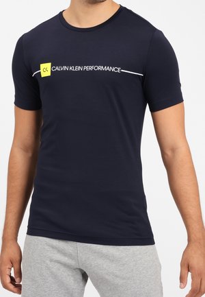 Man die een marineblauw Calvin Klein Performance T-shirt met een geel vierkant logo draagt en lichtgrijze korte broek, staand tegen een witte achtergrond.
