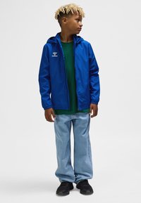 Blå letvægtsjakke med hætte, grøn t-shirt under, løse lyseblå jeans og sorte sneakers. Jakken har logo.