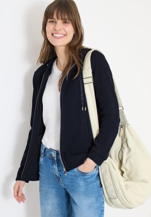 Femme souriante aux cheveux bruns portant une veste noire zippée, un chemisier blanc, un jean bleu, et portant un grand sac à bandoulière beige clair.