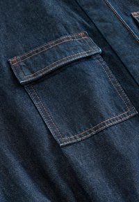 Gros plan sur un tissu denim bleu foncé avec une poche rectangulaire à rabat et des coutures orange visibles.