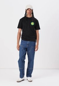 Black cotton t-shirt featuring a circular green graphic. Paired with loose-fitting blue denim jeans and white sneakers. Light-colored cap worn.