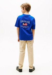 Blaue Baumwoll-T-Shirt mit einem großen weißen "HILFIGER"-Logo, Flaggen-Design und Text auf der Rückseite. Kombiniert mit beigen Hosen und schwarzen Schuhen.