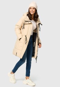 Beige lange pufferjas met een capuchon, zwarte ritsdetails, aantrekkoorden en zakken; gecombineerd met blauwe jeans en witte sneakers.