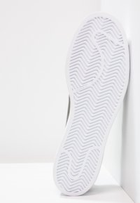Bílá podrážka tenisek s texturovaným cik-cak vzorem, s šedým svrškem viditelným na okraji. Čistý design, moderní materiál, minimalistický styl.