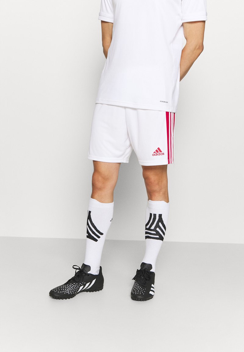 Weißes Sportshirt, weiße Shorts mit roten Adidas-Streifen, schwarze strukturierte Schuhe und weiße Socken mit schwarzem Grafikmuster.