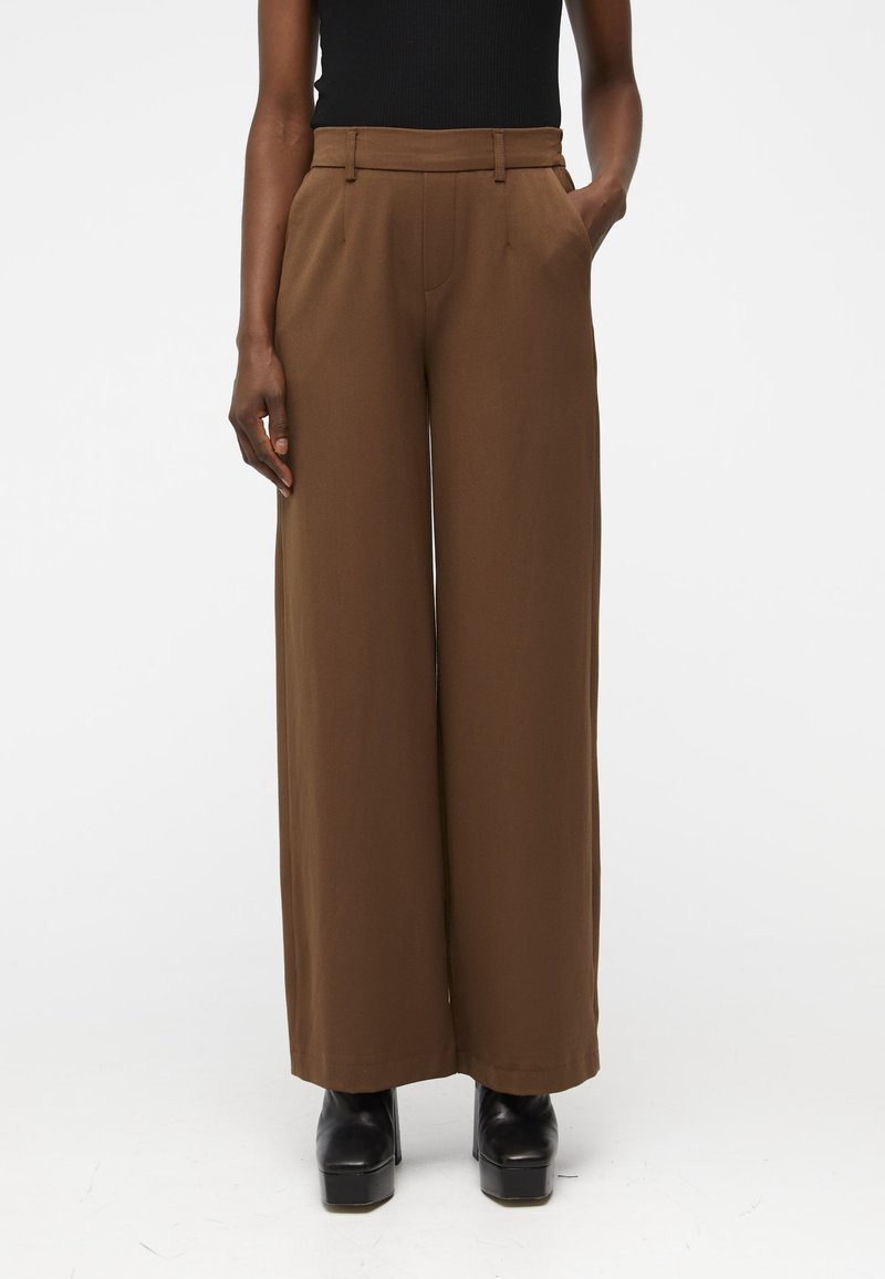 Pantalon large marron à texture lisse, taille haute et poches latérales. Porté avec un haut noir et des chaussures à plateforme noires.