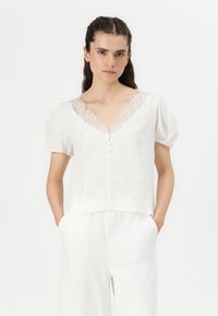 Hvit bluse med V-hals, blondering og puffede ermer. Har en knapp-fra-front og et lett, teksturert stoff.